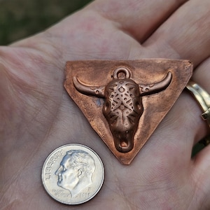 May include: Copper pendant in the shape of a bull's head with intricate detailing, set on a triangular base. A U.S. dime is next to the pendant for size comparison. The pendant has a loop for hanging and is likely a piece of jewelry.