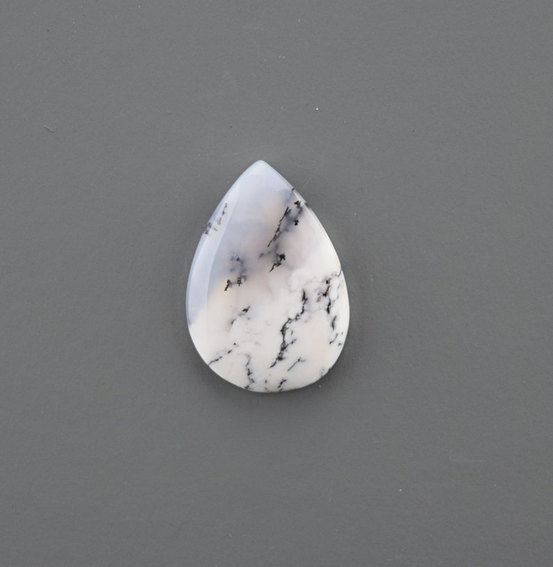 Op de afbeelding: Een traanvormige cabochon van witte dendritische opaal met zwarte dendrieten. De steen is doorschijnend en heeft een melkachtig witte kleur.