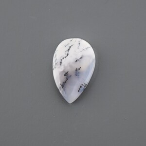 Op de afbeelding: Een traanvormige cabochon van witte opaal met zwarte dendritische insluitsels. De opaal is doorschijnend en heeft een melkachtig witte kleur.