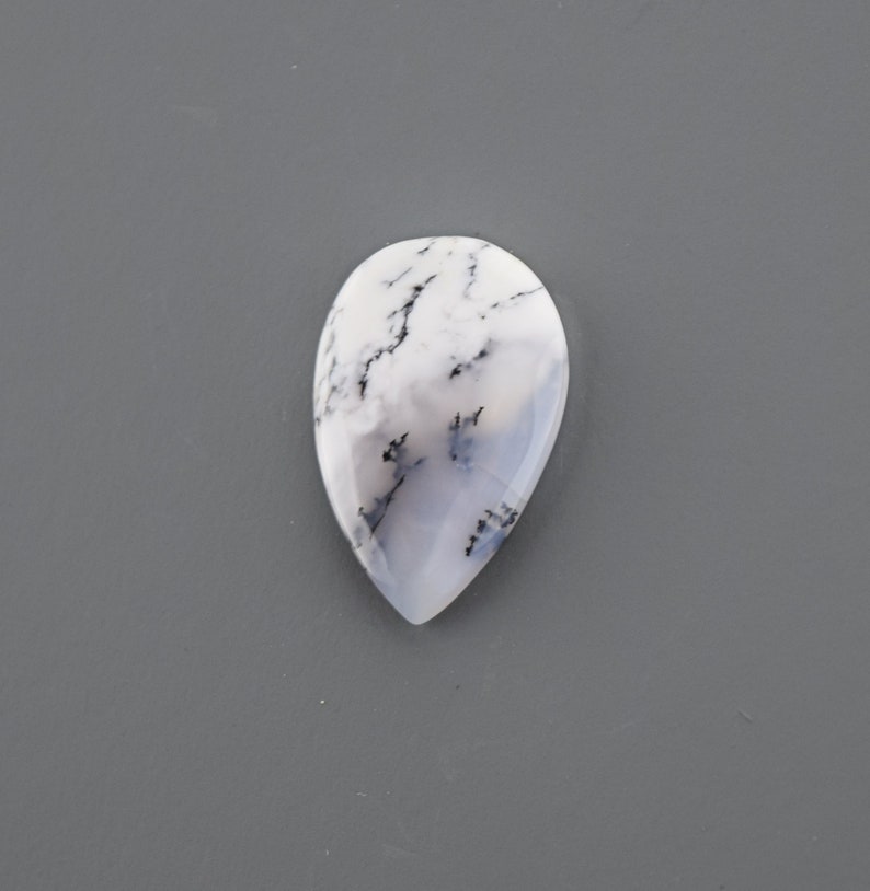 Op de afbeelding: Een traanvormige cabochon van witte opaal met zwarte dendritische insluitsels. De opaal is doorschijnend en heeft een melkachtig witte kleur.