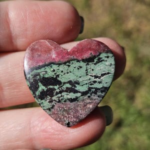 May include: A heart-shaped stone with a polished surface. The stone features bands of red, green, and black. The heart is held in a hand. The text "Littleredjewelrydesign" is visible at the bottom.