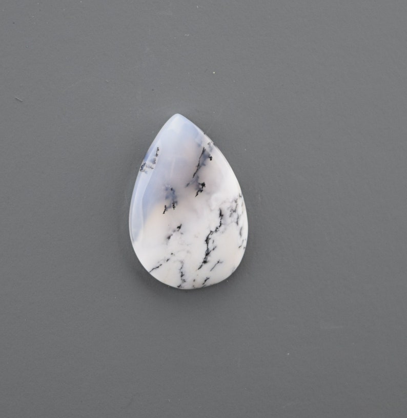 Op de afbeelding: Een witte en grijze traanvormige cabochon met zwarte dendritische insluitsels. De cabochon is een natuursteen met een glad, gepolijst oppervlak.