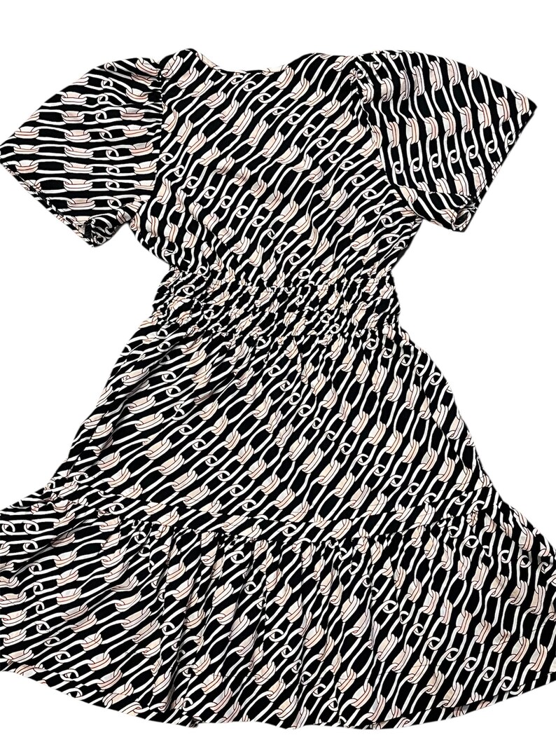 Puede incluir: Vestido estampado en blanco y negro con mangas cortas y cintura fruncida. El vestido presenta un dise&ntilde;o de eslabones de cadena en blanco y beige sobre un fondo negro. La falda tiene un detalle de volantes en capas.