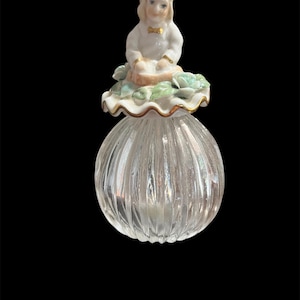 May include: A vintage perfume bottle with a clear, ribbed glass body and a decorative porcelain stopper. The stopper features a seated figure in white clothing with a gold bow tie, surrounded by green and white floral accents.
