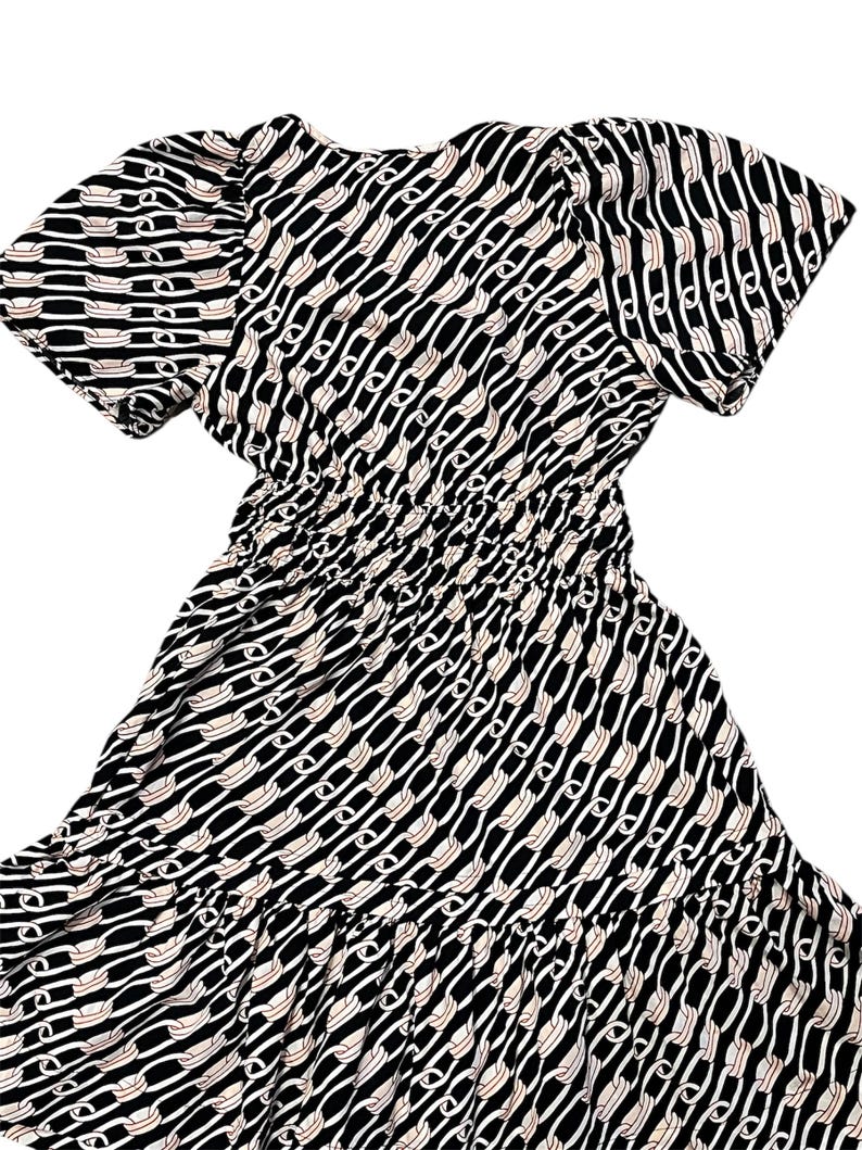 Puede incluir: Vestido estampado en blanco y negro con mangas cortas y cintura fruncida. El vestido presenta un dise&ntilde;o de eslabones de cadena repetidos en blanco y beige sobre un fondo negro. La falda tiene un dobladillo con volantes.