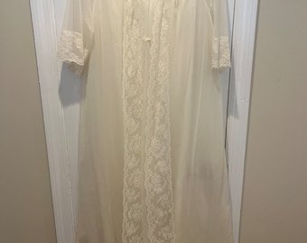 May include: Ivory-colored vintage nightgown with lace trim. The sheer fabric features a delicate lace collar, cuffs, and a vertical lace panel down the front. The nightgown is hanging on a wooden hanger against a white door.