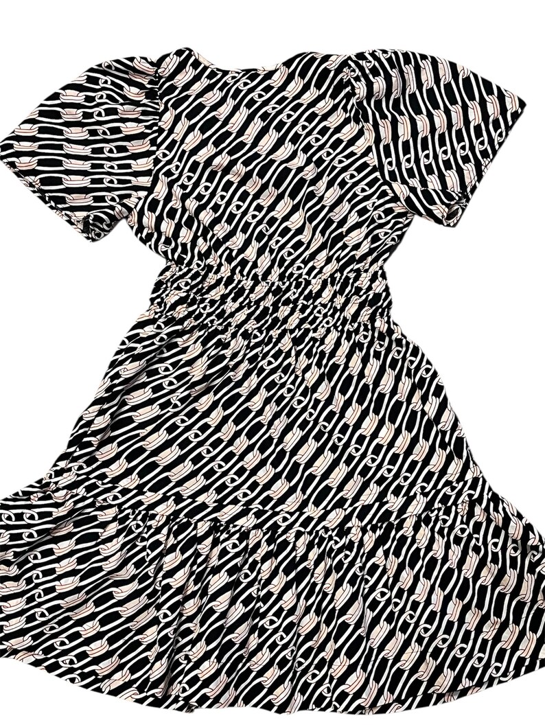 Puede incluir: Vestido estampado en blanco y negro con mangas cortas y cintura fruncida. El vestido presenta un dise&ntilde;o de eslabones de cadena repetidos en blanco y beige sobre un fondo negro. La falda tiene un dobladillo con volantes.