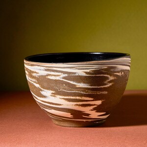 May include: A handcrafted ceramic bowl with a unique marbled design. The bowl features a dark brown exterior with white and tan swirls, and a black interior. The bowl's shape is round with a slightly flared rim, perfect for serving or display.