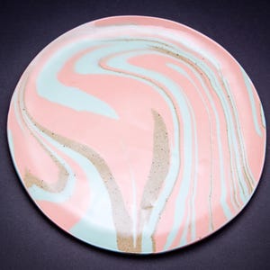 May include: A round ceramic plate with a swirled design in shades of pink, light blue, and tan. The plate has a glossy finish and a smooth surface. The design resembles marble or flowing paint. The plate is a decorative item.