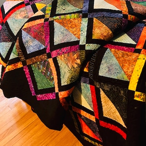 May include: A colorful handmade quilt with a geometric design. The quilt features a patchwork of triangles and squares in various colors, including blue, orange, pink, and green, framed by black borders. The quilt is folded, showing its texture and pattern.