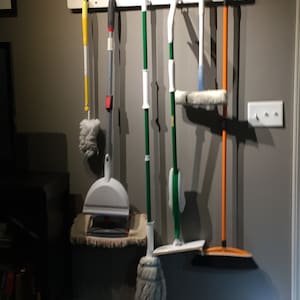 May include: A white wall-mounted rack with six cleaning tools hanging from it. The tools include a yellow duster, a white dustpan, a green mop, a blue squeegee, a green mop with a white head, and an orange broom.