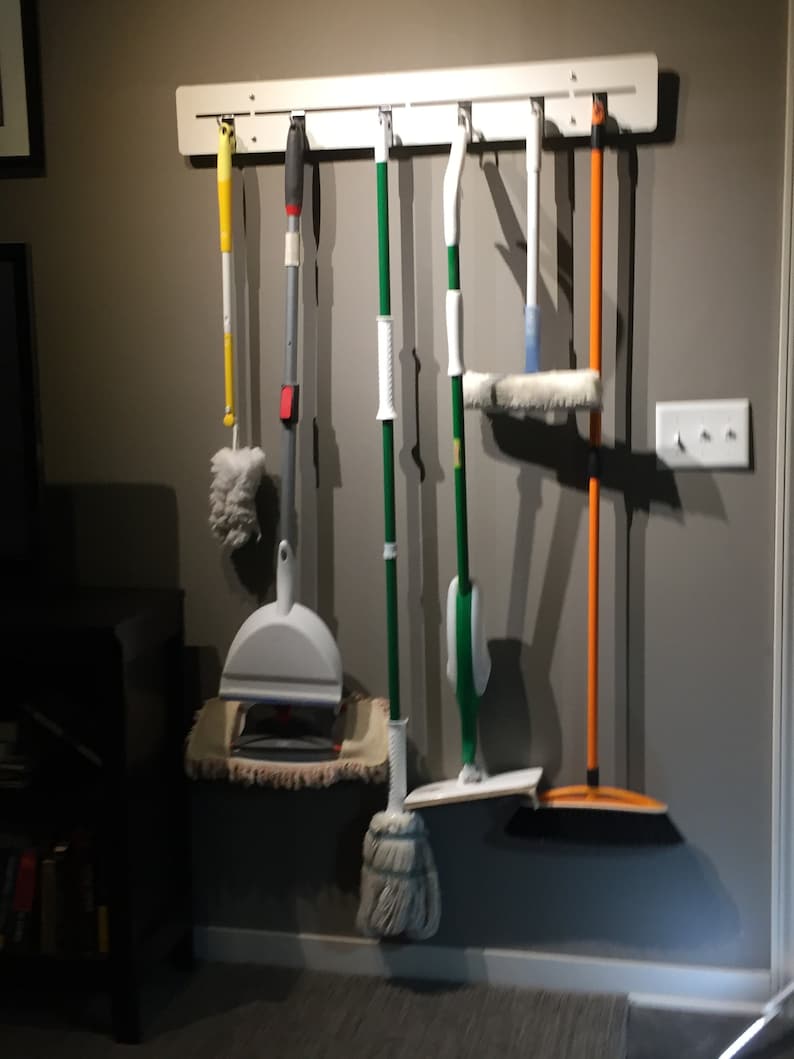 May include: A white wall-mounted rack with six cleaning tools hanging from it. The tools include a yellow duster, a white dustpan, a green mop, a blue squeegee, a green mop with a white head, and an orange broom.