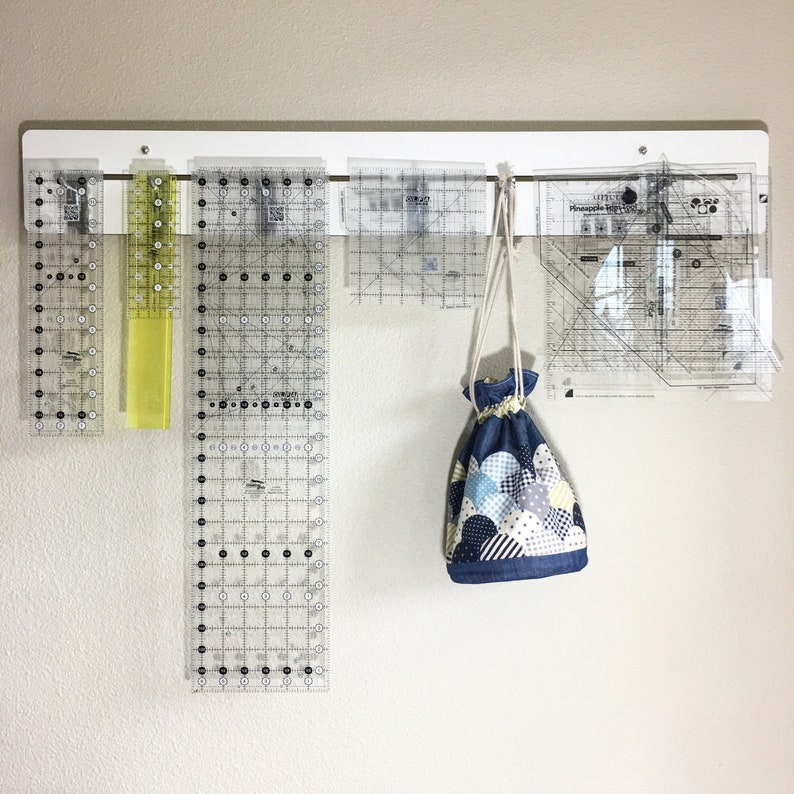 May include: A white wall with a white metal rack holding several clear plastic rulers and a blue and white fabric bag hanging from the rack.