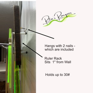May include: A wooden ruler rack with two nails for hanging. The rack sits 1 inch from the wall and can hold up to 30 rulers. The text "Rita's Pacques" is written in green cursive on a white background.