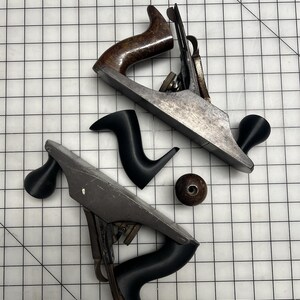 May include: Two metal hand planes with adjustable blades and handles. One plane features a dark brown wooden handle and knob, while the other has matte black handles. A separate small, round, dark brown wooden knob is positioned between them.
