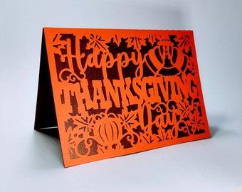 Könnte beinhalten: Orangefarbene Karte mit einem ausgeschnittenen Design mit den Worten "Happy Thanksgiving Day" und Bildern von Truthähnen, Kürbissen und Blättern.