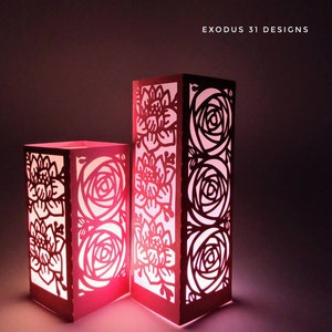 May include: Two illuminated paper lanterns with floral cutouts. The lanterns are rectangular and stand upright, with a warm pink glow emanating from within. The cutouts feature rose and other floral designs. The text "EXODUS 31 DESIGNS" is visible.