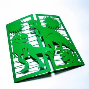 May include: Green paper card with a cut-out design of two dinosaurs in a jungle setting. The card is folded in half and the dinosaurs are visible on both sides.