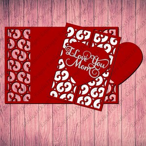 May include: Red and white paper card with a heart-shaped cutout. The heart has the words "I Love You Mom" inside. The card has a decorative border with a repeating pattern.
