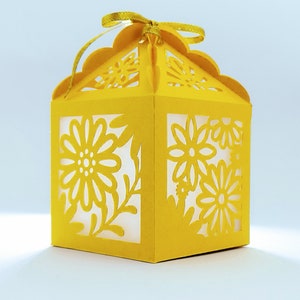May include: A yellow paper gift box with a scalloped top and a floral design. The box has a ribbon tied around it.