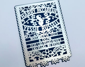 May include: Silver wedding invitation with a blue background and intricate cut-out design. The invitation reads "Harry McCarran & Yessie Russell, November 18, 2023, 6:00 PM, 3131 Las Vegas Blvd, Las Vegas, NV 89109."