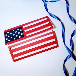 May include: A red, white, and blue paper American flag with a blue ribbon on a white background.