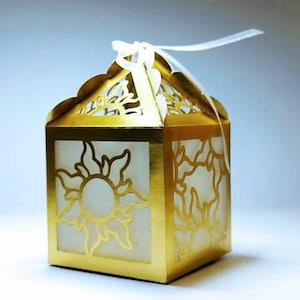 Puede incluir: Una caja de regalo dorada con un diseño de sol blanco y una cinta blanca. La caja está hecha de papel y tiene un diseño delicado e intrincado.