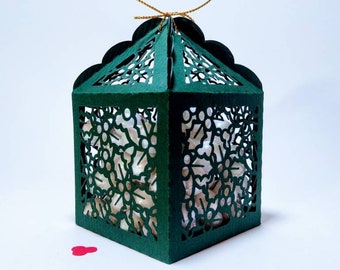 May include: A green paper gift box with a delicate, intricate design of holly leaves and berries. The box is open and has a white paper lining. The box is tied with a gold ribbon.