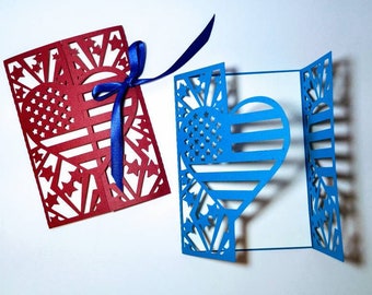 May include: Two paper cards with a heart-shaped cutout in the center. The heart is decorated with the American flag design. One card is red and the other is blue. Both cards are folded in half.