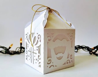 May include: White gift box with a gold ribbon and a cut-out design of a Santa Claus face. The box is sitting on a white surface with a string of Christmas lights in the background.