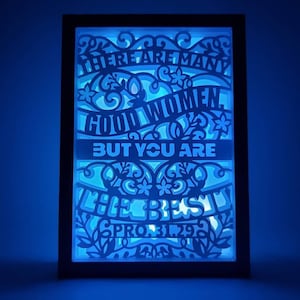 Puede incluir: Una pieza de arte en papel en capas enmarcada con retroiluminación azul. El texto dice "THERE ARE MANY GOOD WOMEN, BUT YOU ARE THE BEST PROV. 31:29" en una fuente decorativa. La obra de arte presenta recortes florales y de desplazamiento.