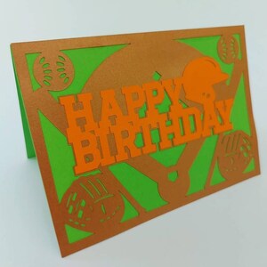 May include: A green and orange paper card with a baseball theme. The card says "Happy Birthday" in orange letters. There are baseballs and a baseball helmet cut out of the card.