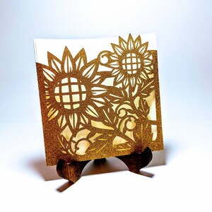 May include: A gold glitter card with a cut-out design of sunflowers and leaves. The card is displayed on a small wooden easel.