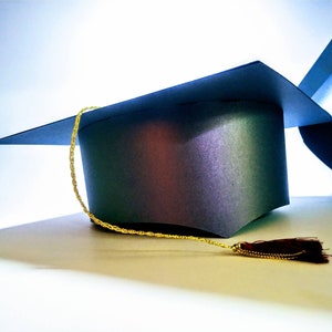 Puede incluir: Dos gorros de graduaci&oacute;n azules con un borla dorada y una borla roja. Los gorros est&aacute;n sobre una superficie blanca.