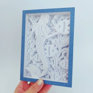 May include: A white paper cutout of a mermaid with a blue frame. The mermaid is surrounded by fish and seaweed.