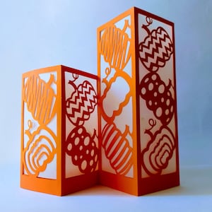 Op de afbeelding: Twee oranje papieren lantaarns met uitgesneden pompoenontwerpen. De lantaarns zijn gemaakt van wit papier met oranje uitsnijdingen van pompoenen met verschillende patronen, waaronder strepen, stippen en zigzag.