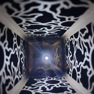 May include: A black and white patterned box with a light shining through the opening. The pattern is made up of repeating sun shapes.