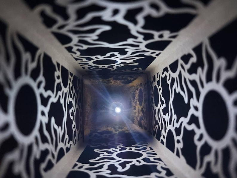 May include: A black and white patterned box with a light shining through the opening. The pattern is made up of repeating sun shapes.