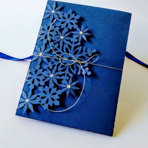 K&ouml;nnte beinhalten: Eine marineblaue Karte mit einem dekorativen Schneeflocken-Design, das aus dem Papier ausgeschnitten ist. Die Schneeflocke ist mit silbernem Glitzer und Strasssteinen verziert.