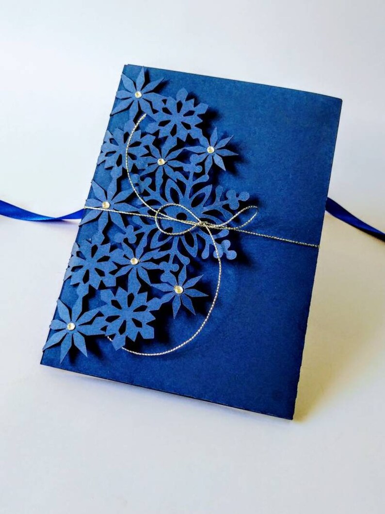 K&ouml;nnte beinhalten: Eine marineblaue Karte mit einem dekorativen Schneeflocken-Design, das aus dem Papier ausgeschnitten ist. Die Schneeflocke ist mit silbernem Glitzer und Strasssteinen verziert.