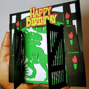 May include: A green dinosaur pops up from behind a black gate with a "Happy Birthday" banner on top. The gate is decorated with red flowers.