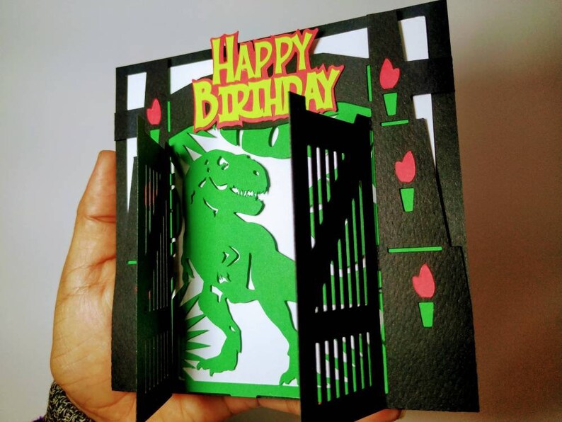 May include: A green dinosaur pops up from behind a black gate with a "Happy Birthday" banner on top. The gate is decorated with red flowers.