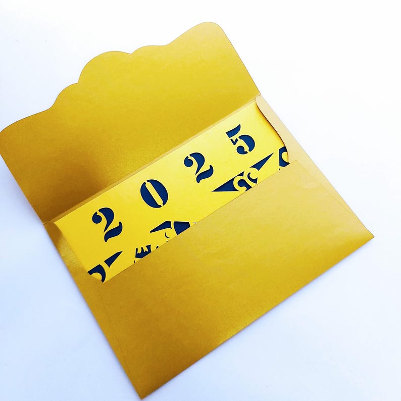 以下が含まれることがあります： 青い2025のカットアウトデザインが施された金の封筒。封筒が開いており、カットアウトデザインが見えています。