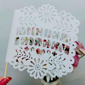 May include: A white paper cutout with the words "Mi Querida" in a decorative font. The cutout has a floral and heart design and is attached to a wooden stick. The background features pink flowers.