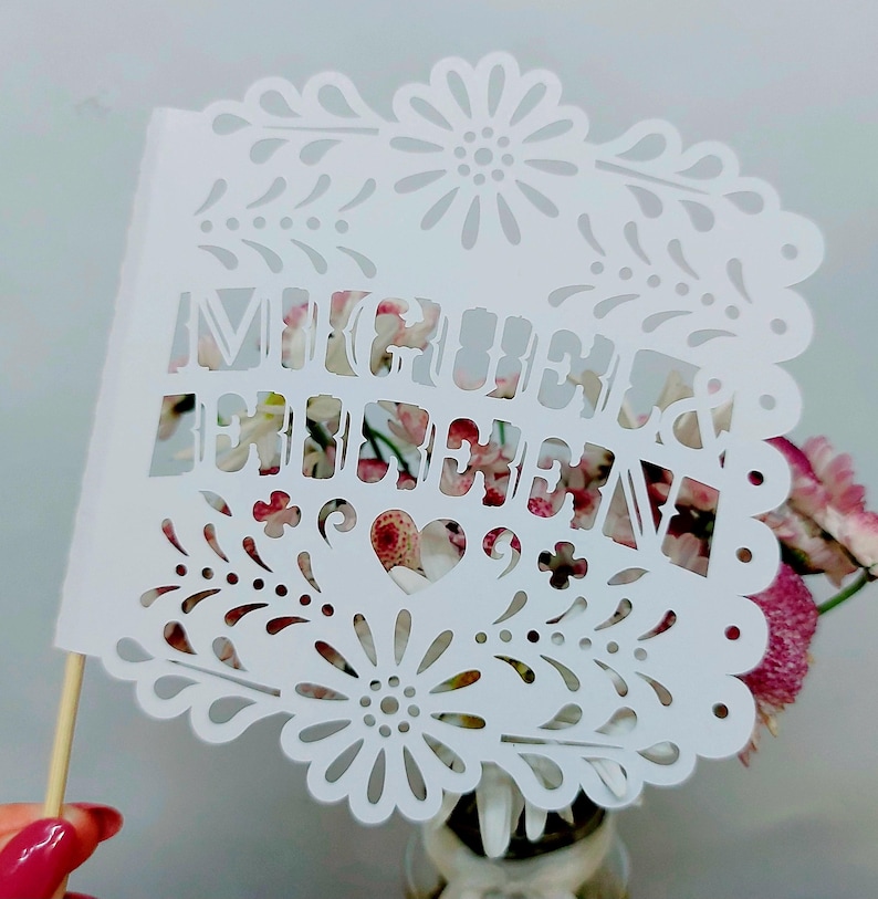 May include: A white paper cutout with the words "Mi Querida" in a decorative font. The cutout has a floral and heart design and is attached to a wooden stick. The background features pink flowers.