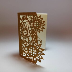 May include: A gold card with a cut-out design of sunflowers and leaves. The card is folded in half and the inside is blank.