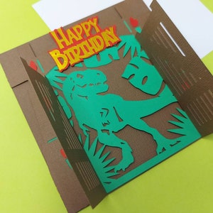 May include: A brown card with a pop-up dinosaur. The dinosaur is green and has a textured brown background. The card says "Happy Birthday" in yellow letters with red outlines.