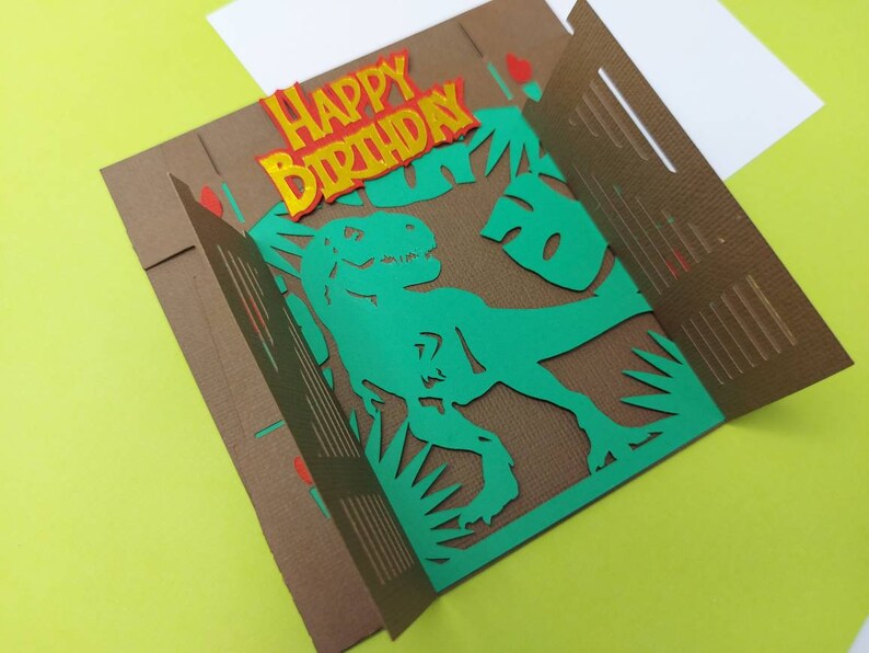May include: A brown card with a pop-up dinosaur. The dinosaur is green and has a textured brown background. The card says "Happy Birthday" in yellow letters with red outlines.