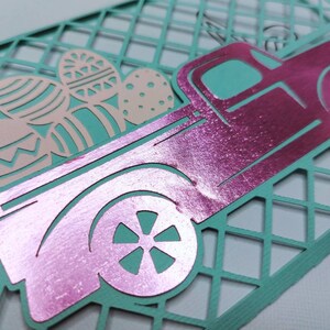 May include: A pink metallic pickup truck carrying pastel pink and blue Easter eggs. The truck is on a teal blue background with a diamond pattern.