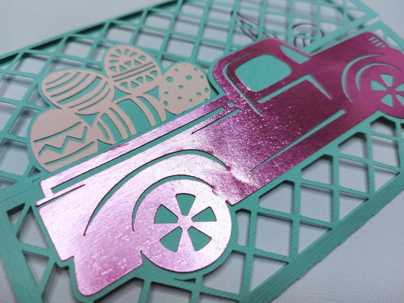 May include: A pink metallic pickup truck carrying pastel pink and blue Easter eggs. The truck is on a teal blue background with a diamond pattern.